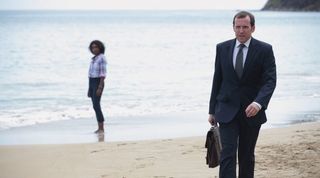 Richard Poole on a beach in Death in Paradise with Camille in the background